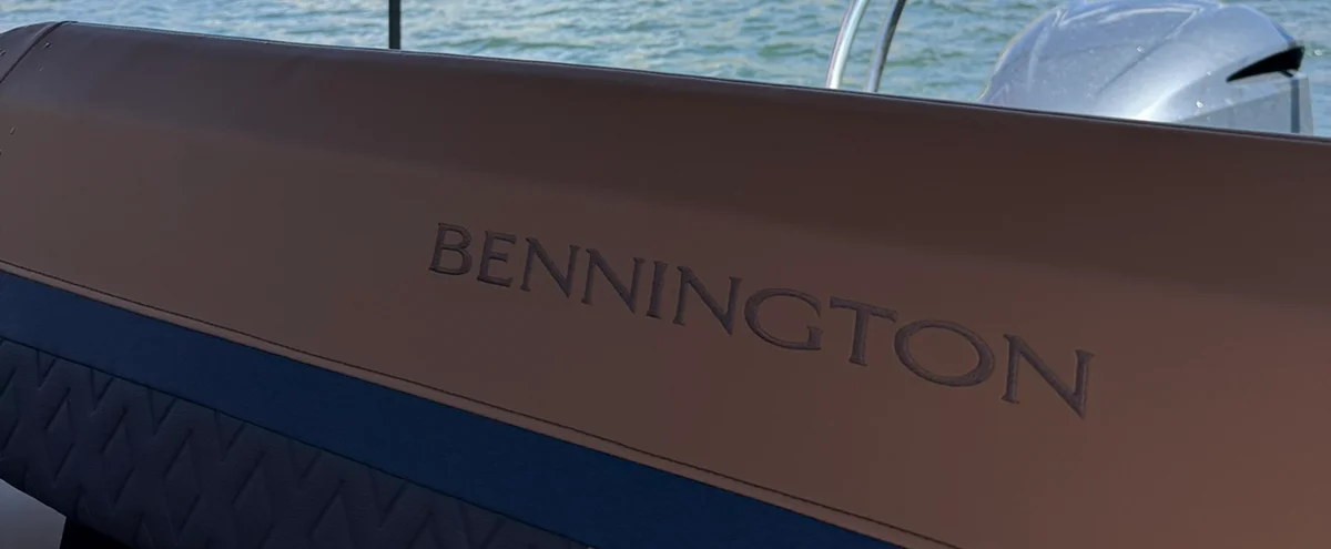 A close-up of the "BENNINGTON" brand name embossed on tan upholstery inside a boat, with a silver outboard motor and water visible in the blurred background