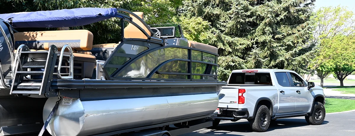 A silver pickup truck towing a Bennington M24 pontoon boat on a trailer along a driveway with trees in the background