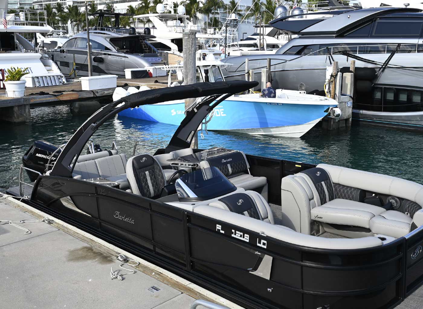 A black Barletta pontoon boat with grey upholstery docked in a marina with several large luxury yachts in the background.