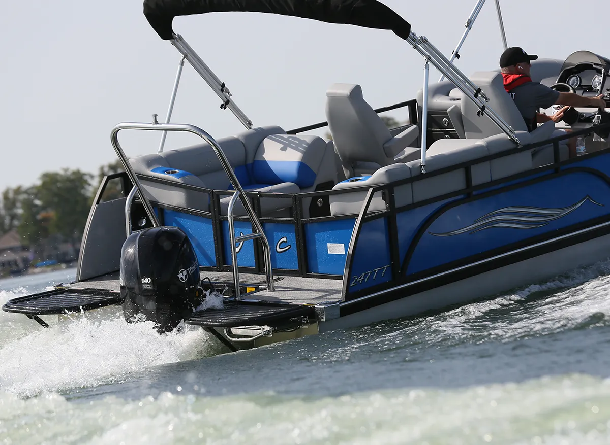 A rear-angle shot of a JC 247TT pontoon boat in motion. The boat features blue side panels, grey and blue upholstery, a black bimini top, and a 140hp Tohatsu outboard motor