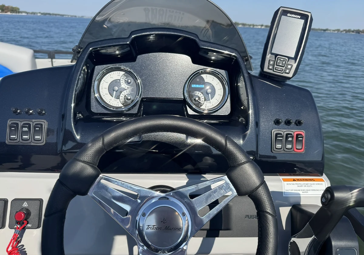 A view of the helm of a JC TriToon Marine boat, featuring a black dashboard with analog gauges, a Garmin GPS unit, and a leather-wrapped steering wheel