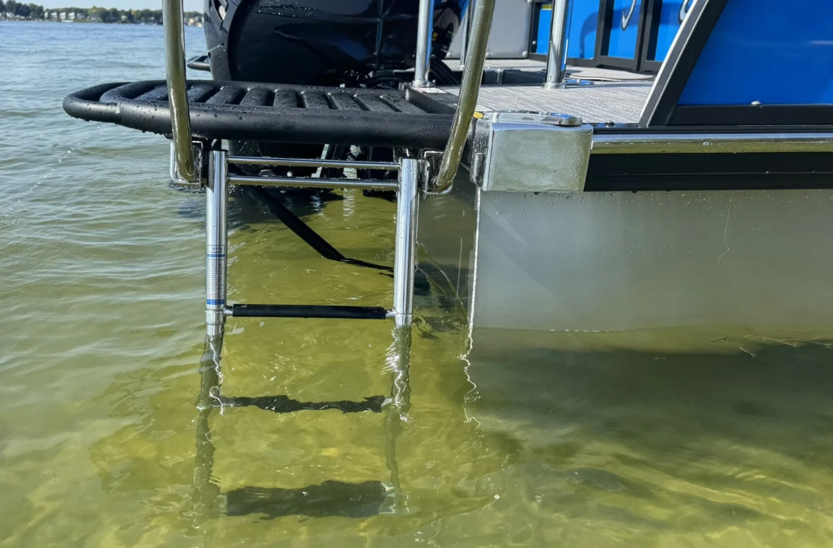 A close-up of a pontoon boat's rear swim platform and a stainless steel boarding ladder extending into shallow water