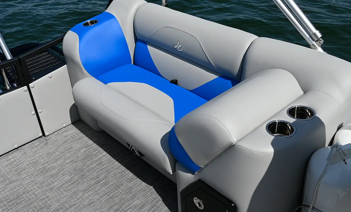 A close-up of the grey and blue lounge seating on a JC Spirit pontoon boat, featuring two integrated stainless steel cup holders and an embroidered "JC" logo on the backrest