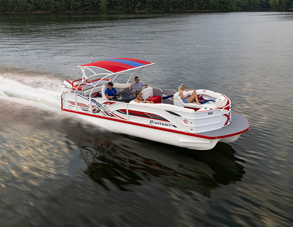 A white PlayCraft pontoon boat with red and black graphics and a red and blue bimini top cruises on a lake, creating a white wake. Four people are on board. A treeline is visible in the background across the calm water.
