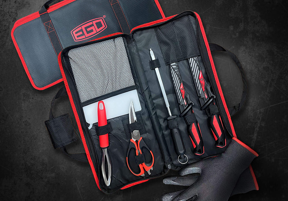 Open black and red EGO TIDEWATER Tool Kit case containing a fish scaler, scissors, cutting board, sharpening rod, and two fillet knives. A pair of black PVC-coated fishing gloves sits next to the case on a dark surface.