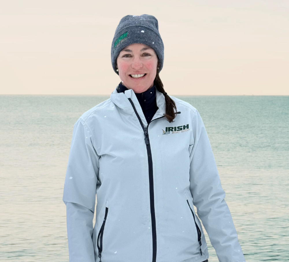 A smiling woman in a gray beanie and a light gray zip-up jacket with the Irish Boat Shop logo stands in front of a calm body of water under a pale sky.
