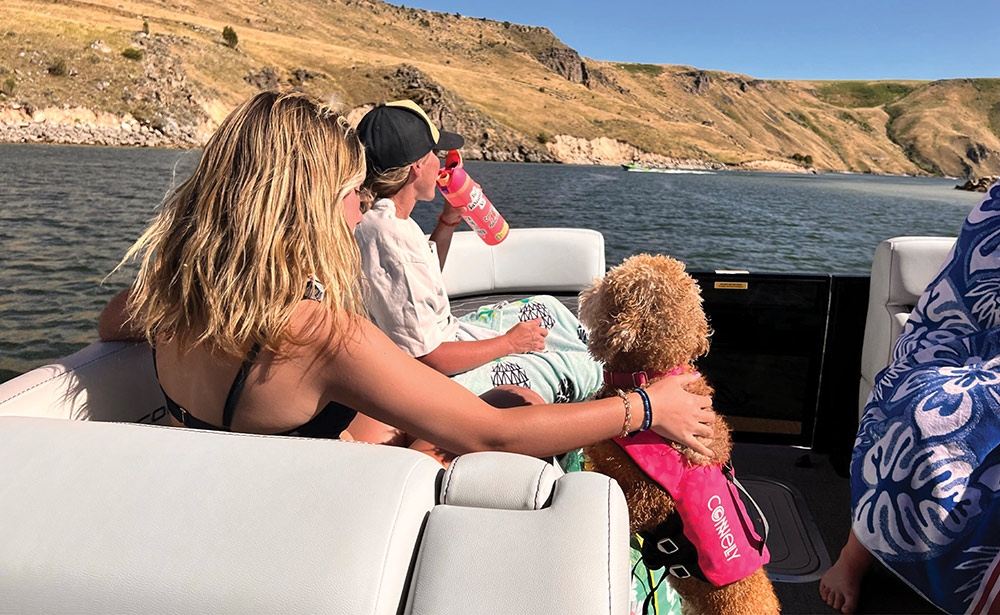 Two people and a dog in a life vest ride on a pontoon boat.