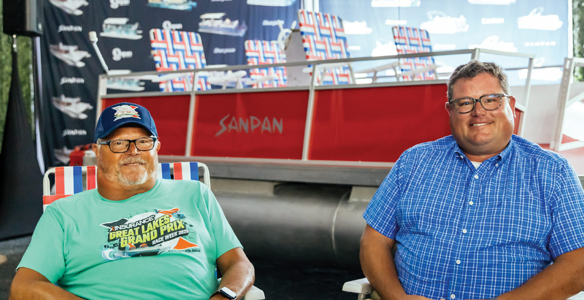 Two men sit in front of a red and silver Sanpan pontoon boat; one wears a green t-shirt and the other a blue plaid shirt.