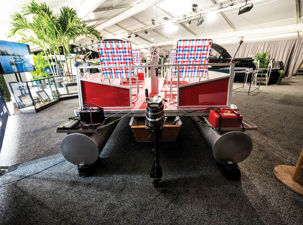 Rear view of a pontoon boat on a carpeted floor, showing the outboard motor, battery, and red fuel tank.