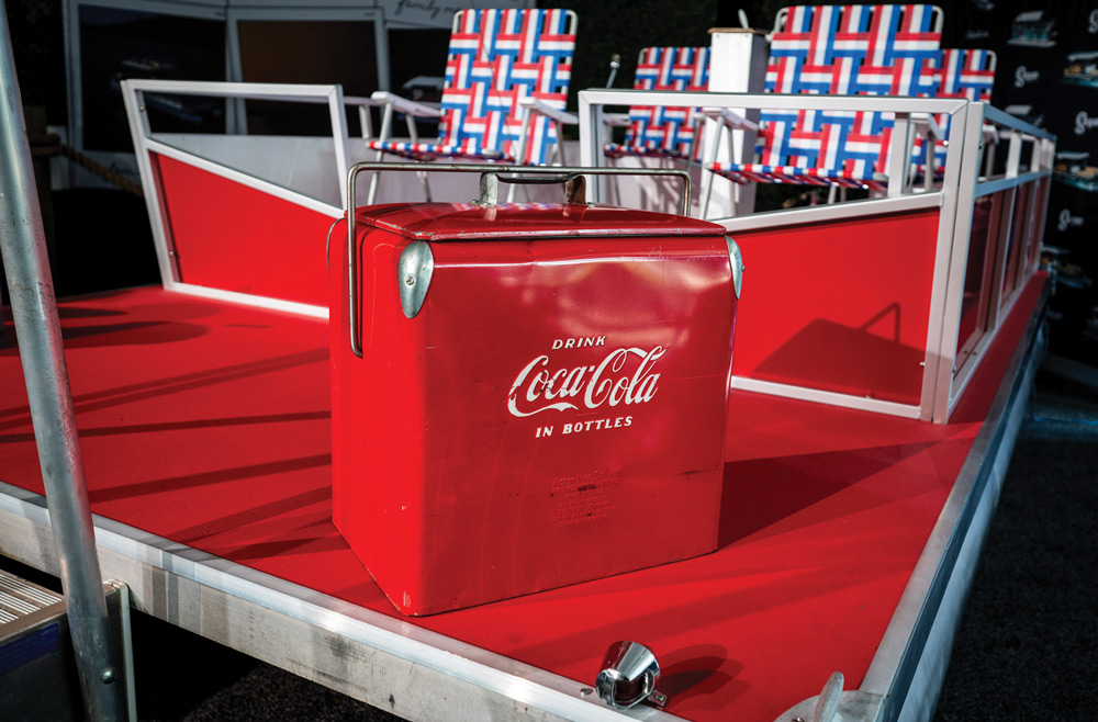 A vintage red metal Coca-Cola cooler sits on the red deck of a boat in front of red, white, and blue woven lawn chairs.