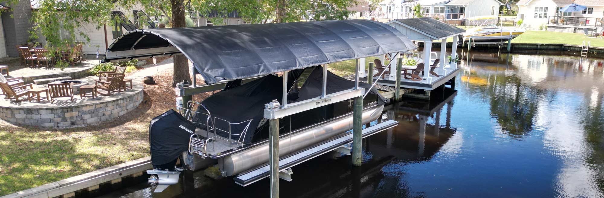 A wide shot of a black pontoon boat lift with a large protective black canopy installed over a dock on a calm waterway.