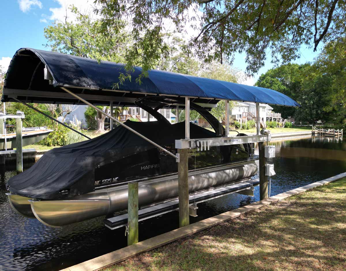 A close-up view of a pontoon boat resting on a lift beneath a curved black Coastal Canopy, situated next to a grassy shoreline.