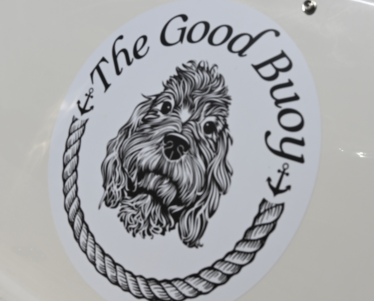 "The Good Buoy" circular decal featuring a black and white illustration of a fluffy dog's head, framed by anchors and a rope border.