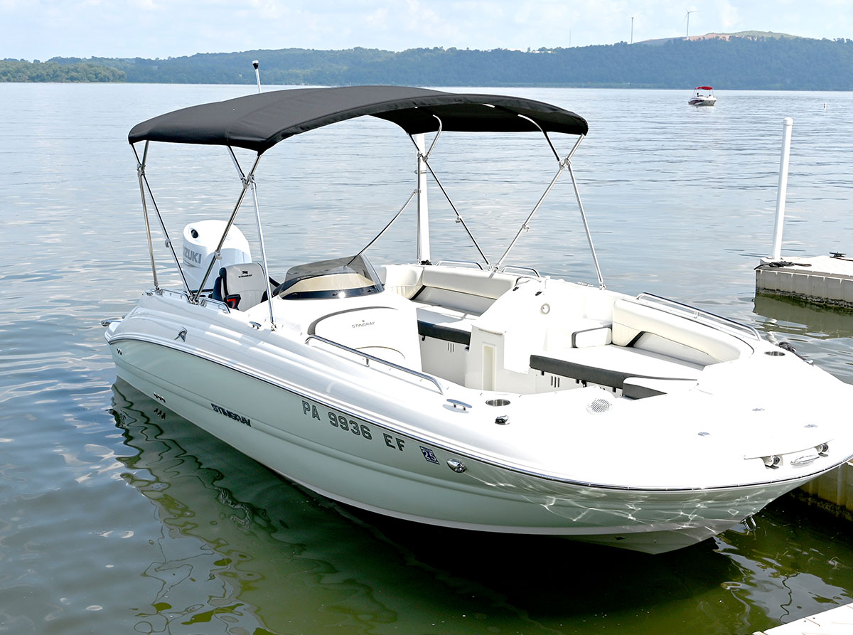 A white Stingray motorboat with a black bimini top and a white Suzuki outboard motor is docked at a pier on a calm lake, with hills visible in the background.