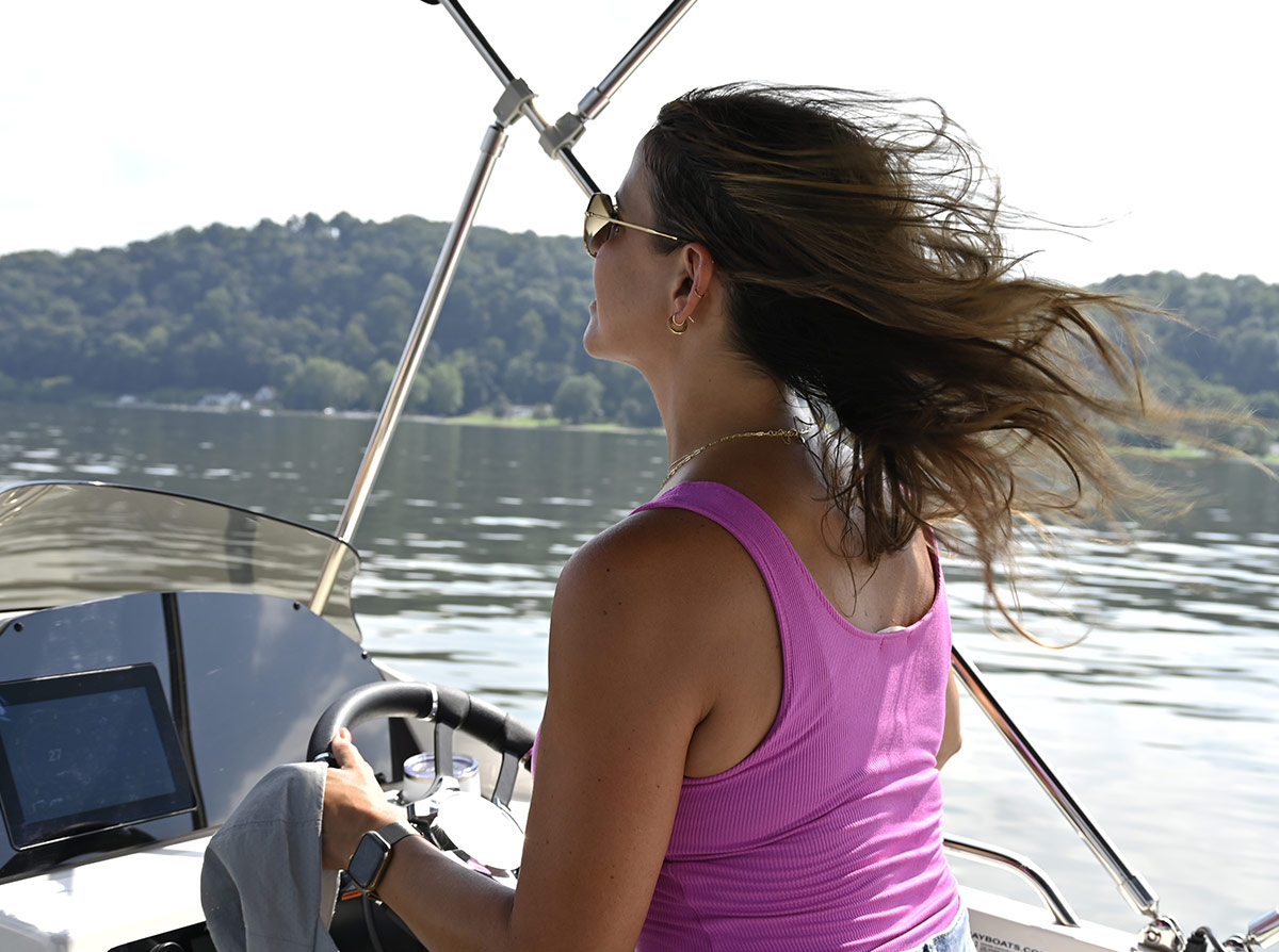 A woman in a pink tank top and sunglasses steers a motorboat on a calm lake, her hair blowing in the wind. A forested hill is visible in the background.