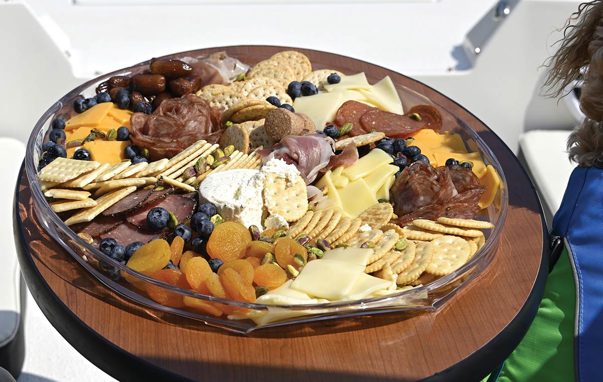 Close-up of a diverse charcuterie spread featuring meats, cheeses, crackers, dried apricots, and berries on a clear tray, served on a boat's wooden table.