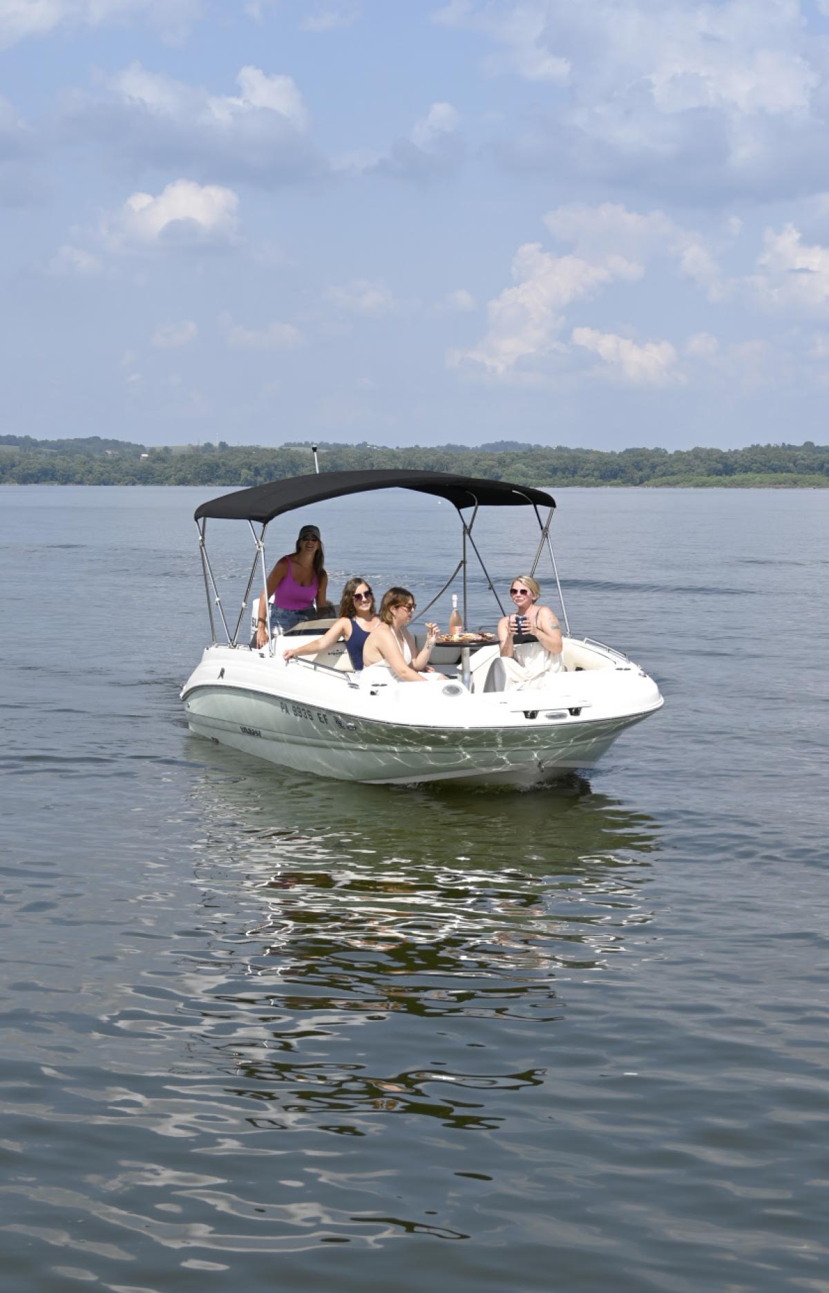 A group of four women enjoys a sunny day on a white deck boat with a black bimini top, cruising on a wide, calm lake.