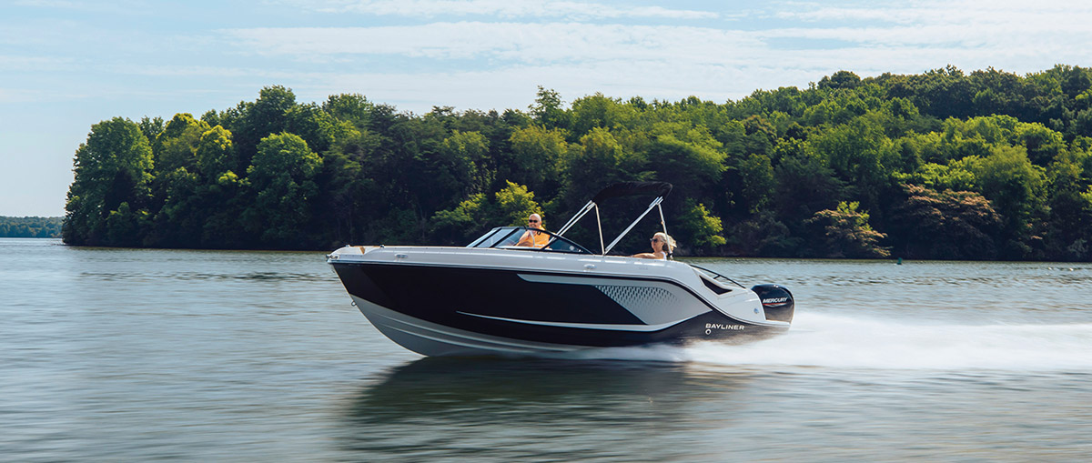 A black and white Bayliner motorboat carrying two people speeds across a lake, with a lush green treeline and blue sky in the background.