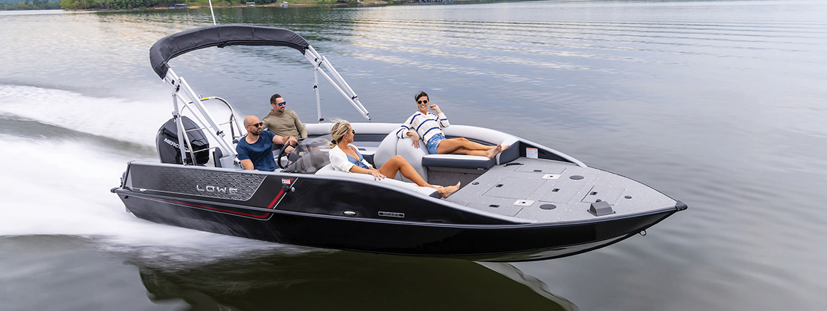 Four people relax on a black and grey Lowe motorboat with a bimini top as it cruises across a calm lake, leaving a white wake.