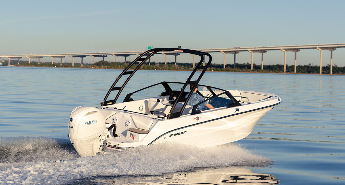 A white Stingray motorboat with a black wake tower and a Yamaha outboard motor speeds across the water. A long bridge is visible in the background under a clear, blue sky.