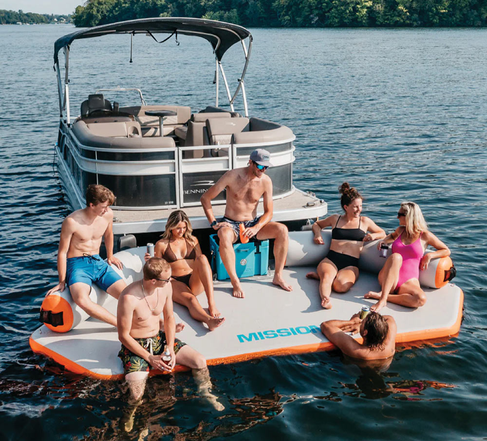 A group of seven young adults relaxing on a large, inflatable grey and orange REEF Deck from Mission Outdoors attached to the back of a pontoon boat