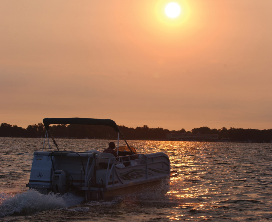 A pontoon boat cruises on a lake during a vibrant orange sunrise or sunset