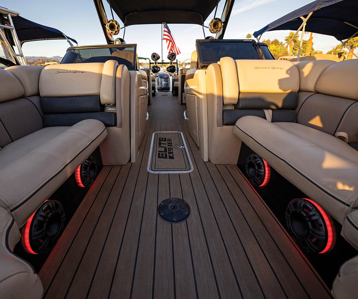 Interior of a South Bay boat featuring tan upholstered seating and wood-grain flooring. Several marine speakers with red LED lighting are integrated into the seat bases, with an "ELITE AUDIO" logo on a floor hatch.