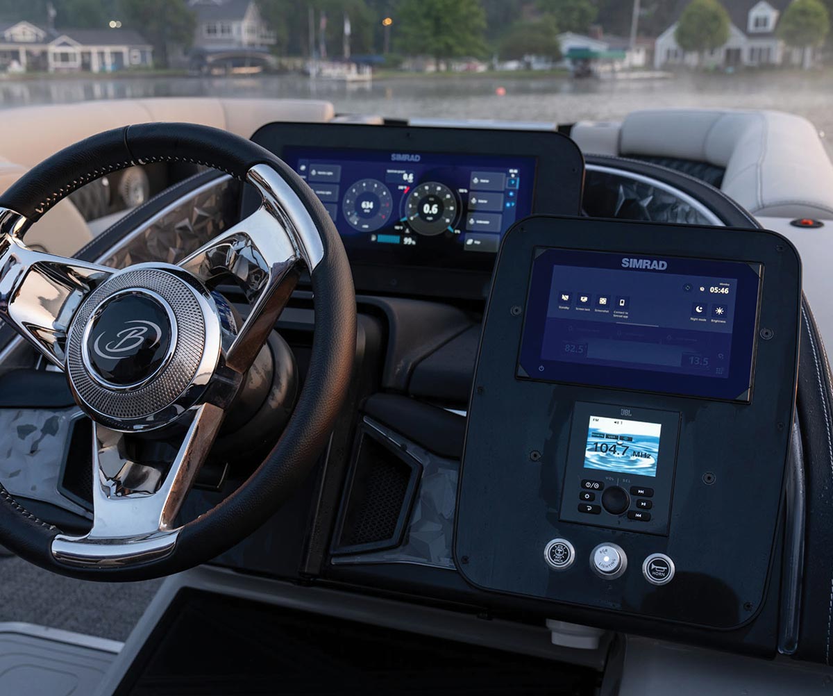 Close-up of a Barletta boat helm featuring a black and silver steering wheel, two Simrad digital displays, and a JBL marine stereo. The dashboard overlooks a calm lake with houses in the background.