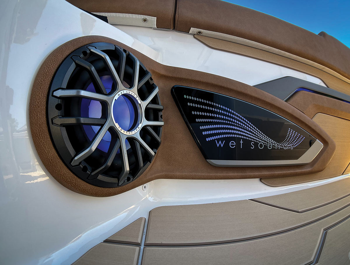 Close-up of a Wet Sounds marine speaker with blue LED lighting and a branded decorative panel, integrated into a boat's white and tan interior with wood-grain flooring.