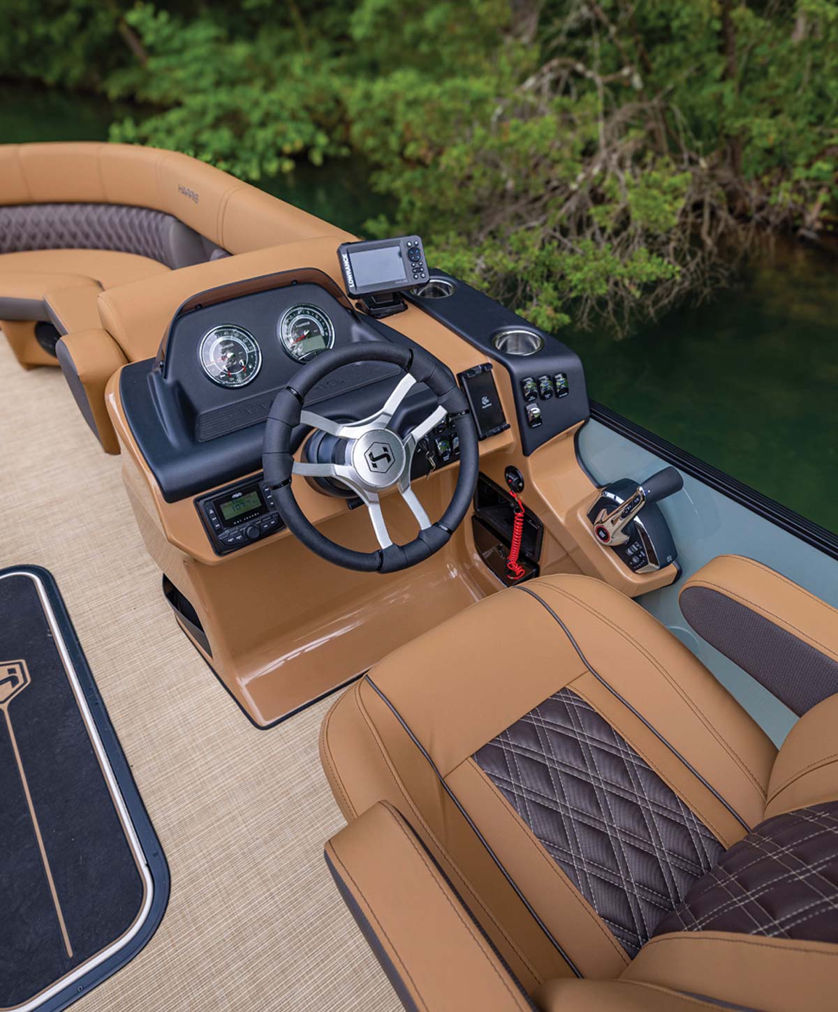 Interior of a Harris pontoon boat helm featuring tan quilted seating, a black and silver steering wheel, and a dashboard with gauges, overlooking calm water and lush green trees.
