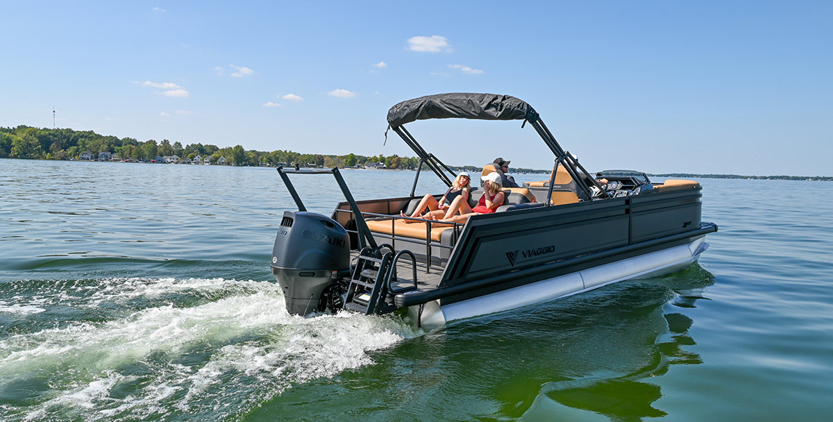 Three people cruise on a black Viaggio pontoon boat with tan seating and a Suzuki outboard motor. The boat leaves a white wake on a calm lake, with a treeline and blue sky in the background.