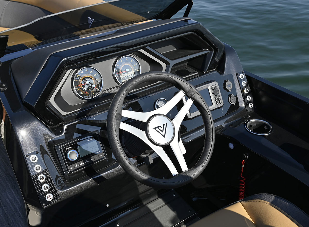Close-up of a Viaggio boat helm featuring a black and silver steering wheel with a "V" logo, two analog gauges, a Clarion stereo, and control switches on a sleek black dashboard next to tan seating.