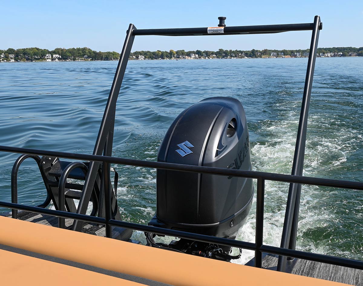 A matte black Suzuki outboard motor on a boat moves through calm water, creating a white wake. The motor is framed by a black tow bar, with a distant shoreline and blue sky in the background.