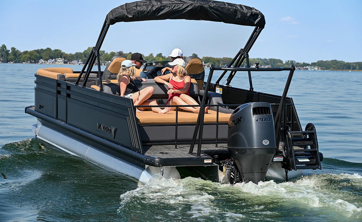 Four people relax on a black Viaggio pontoon boat with tan seating and a Suzuki outboard motor. The boat moves across a calm lake, leaving a white wake under a clear blue sky.