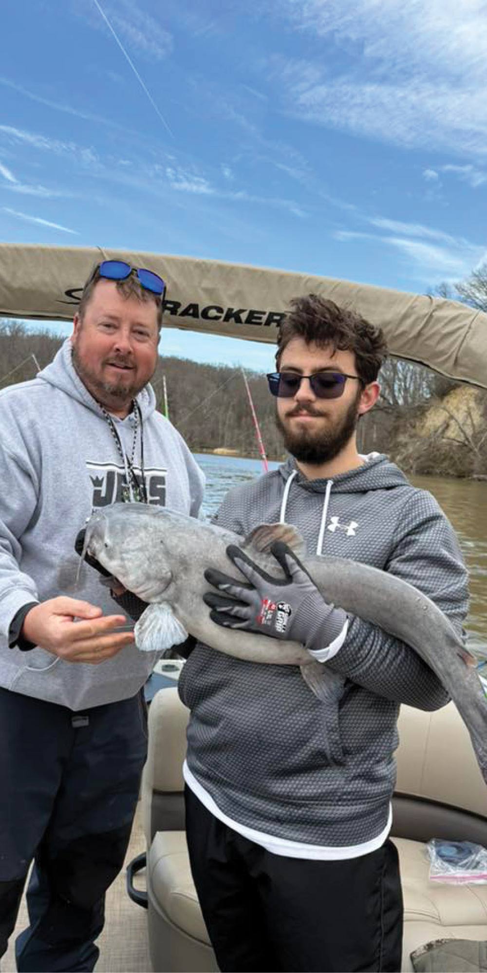Two men standing on a pontoon boat, proudly displaying and holding a large grey catfish