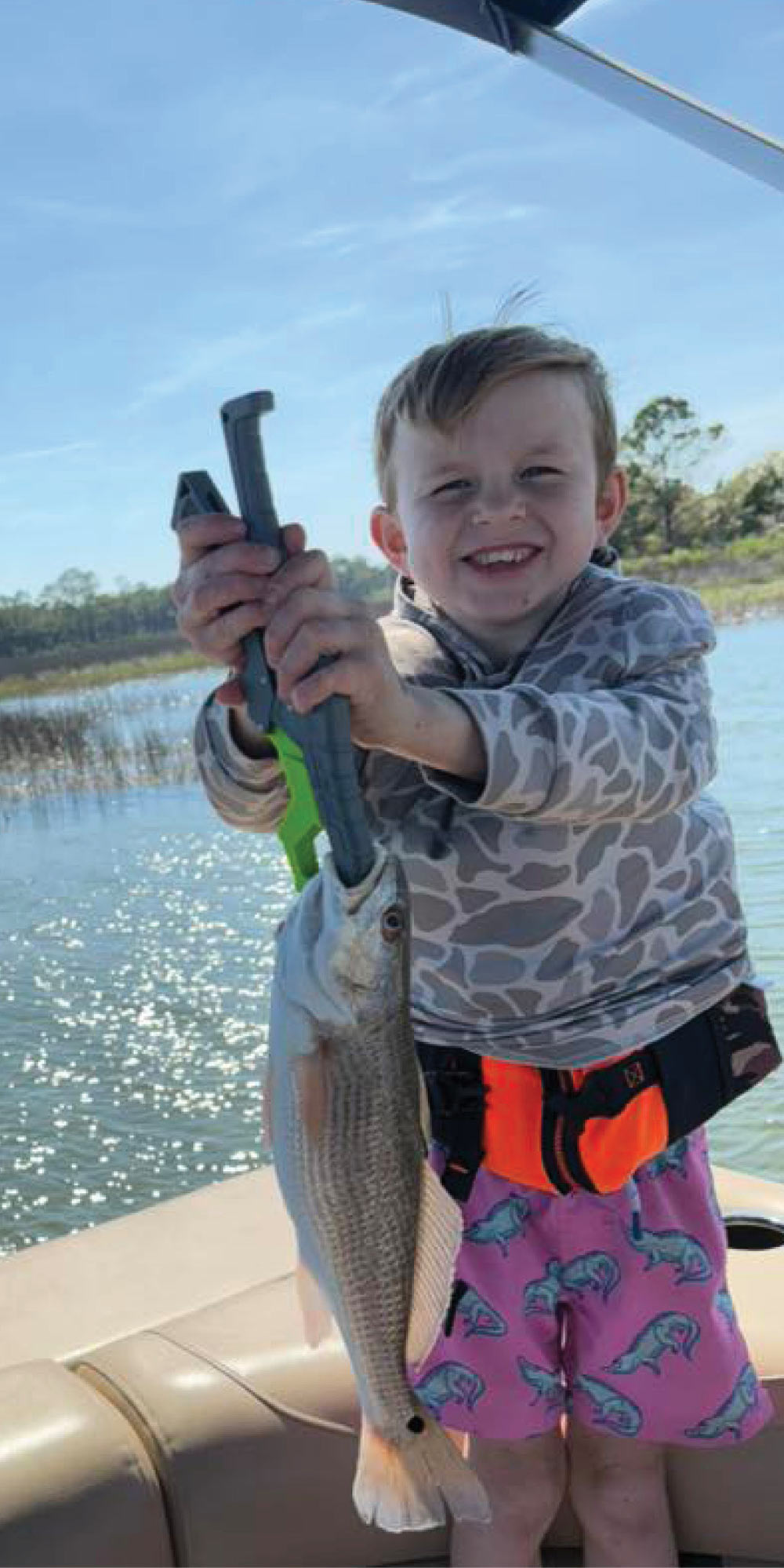 A young boy standing on a pontoon boat with a joyful expression holding up a silvery-tan fish (a Red Drum) using a green and black lip-grip tool