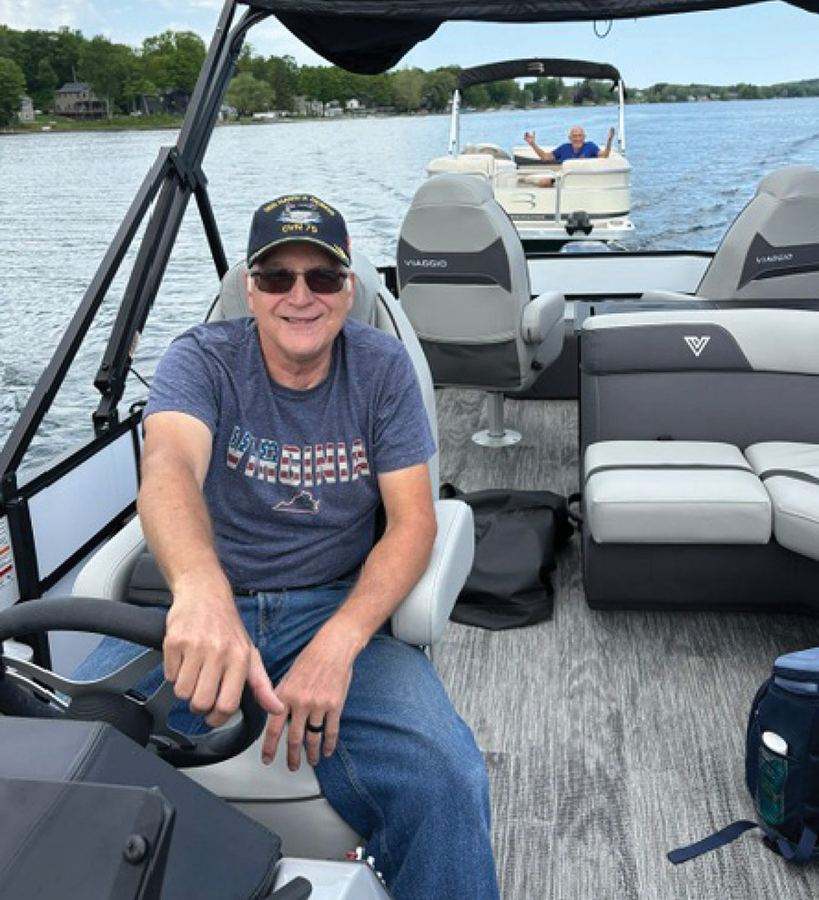 A smiling man in a cap steers a pontoon boat on the water.