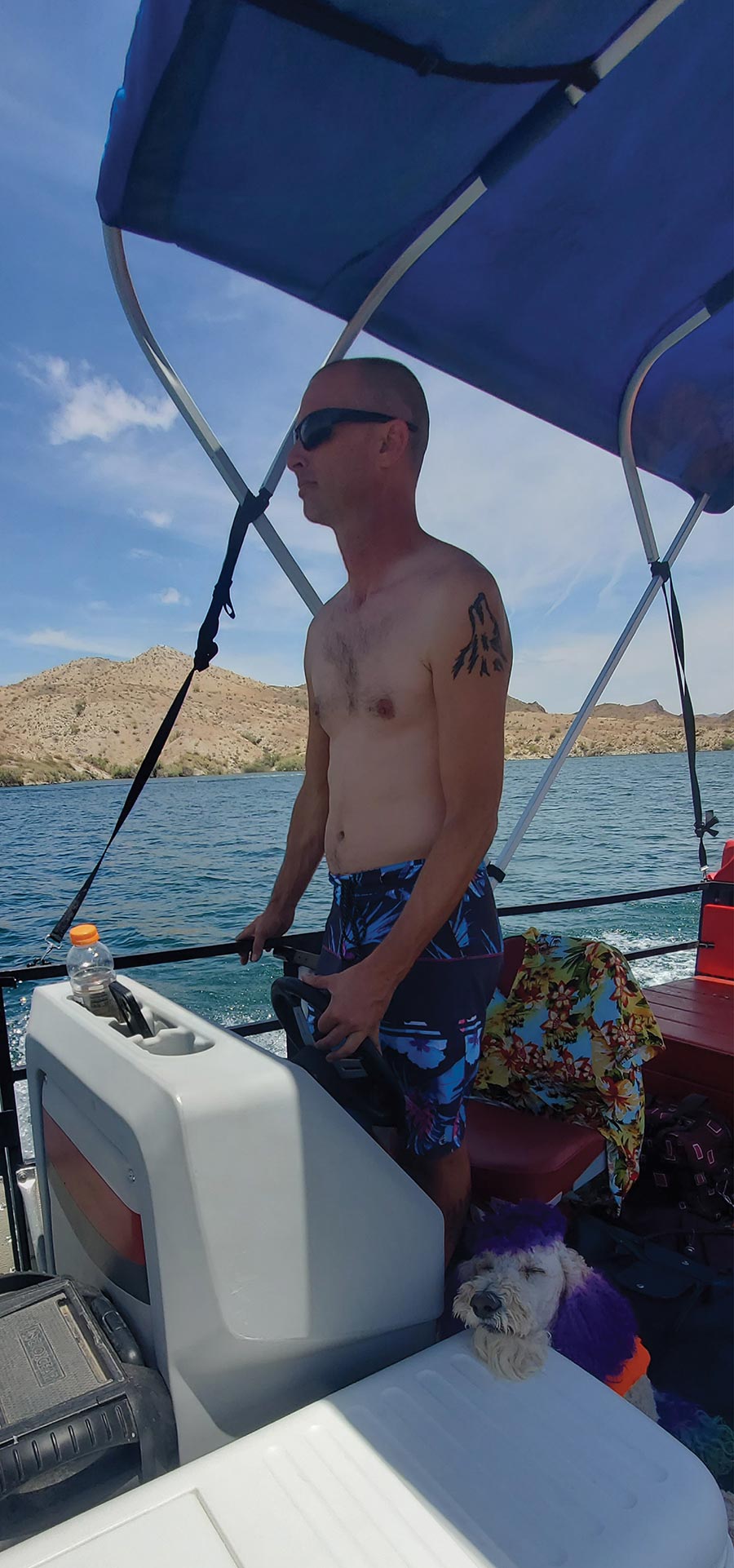 Shirtless man wearing sunglasses steering a pontoon boat on sunny water.