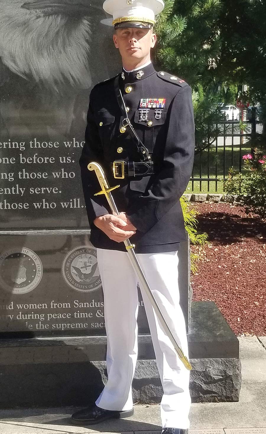 Man in a dress uniform holding a ceremonial sword next to a monument.