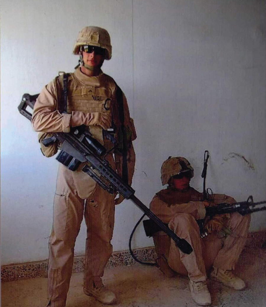 Two soldiers in full combat gear pose with a large sniper rifle indoors.