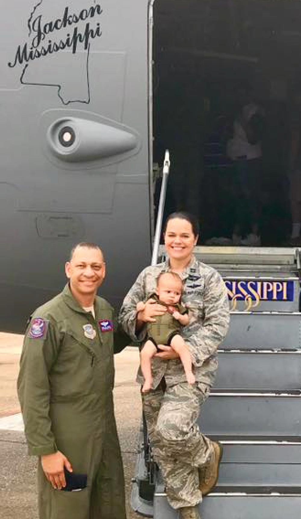 Two smiling military service members stand outside an airplane door holding a baby.