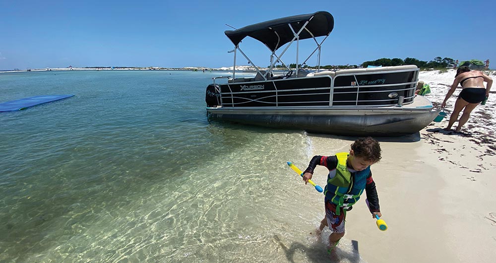 A young child wearing a life jacket plays on the shore near a pontoon boat.