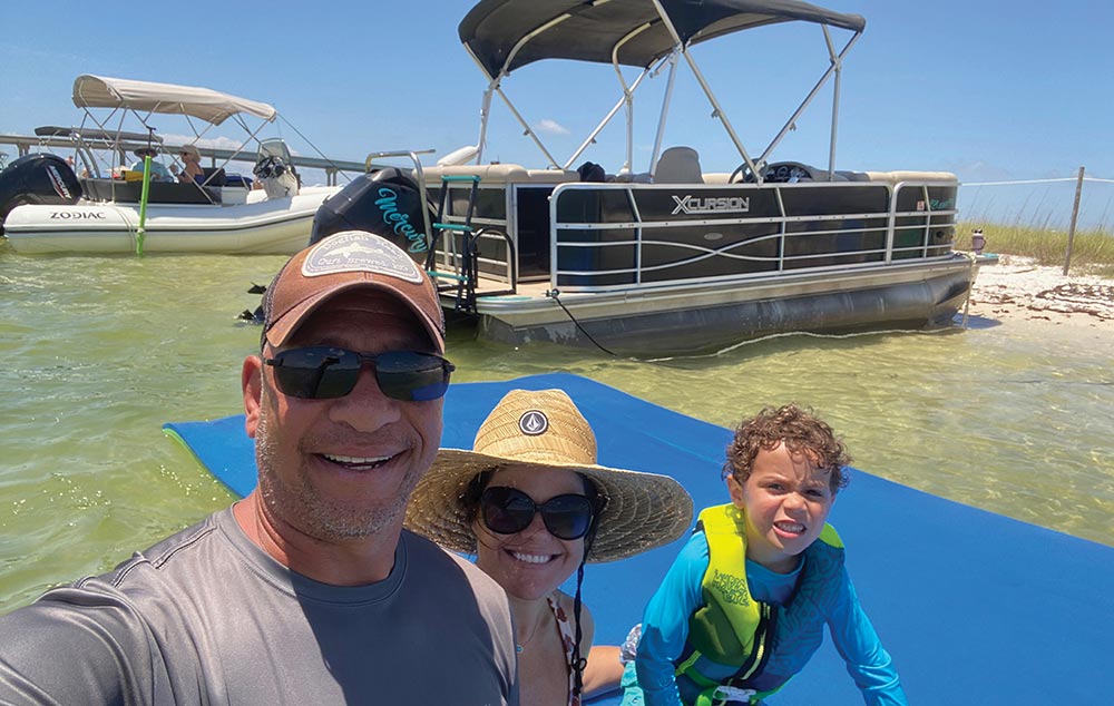 A family smiles while floating on a blue water mat near their pontoon boat.