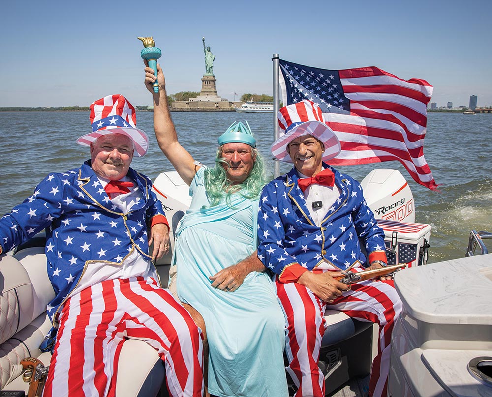 Three smiling men dressed in patriotic United States of America costumes seated on a pontoon boat in the water, with the Statue of Liberty landmark in the background
