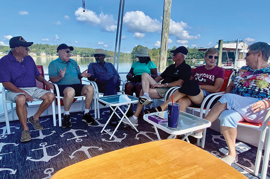 A group of people sit in chairs on a boat deck talking on the water.