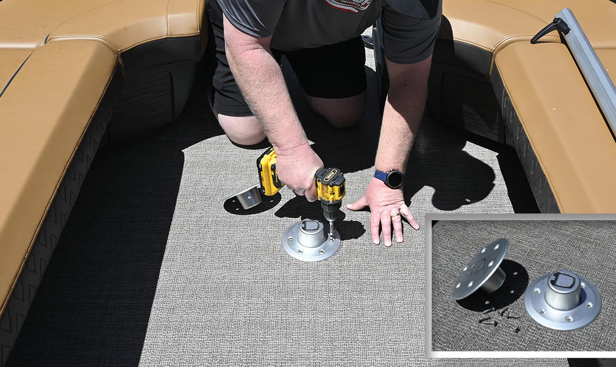 A man kneeling on a boat's grey woven floor uses a yellow cordless drill to install a circular silver metal base plate between tan marine seats.