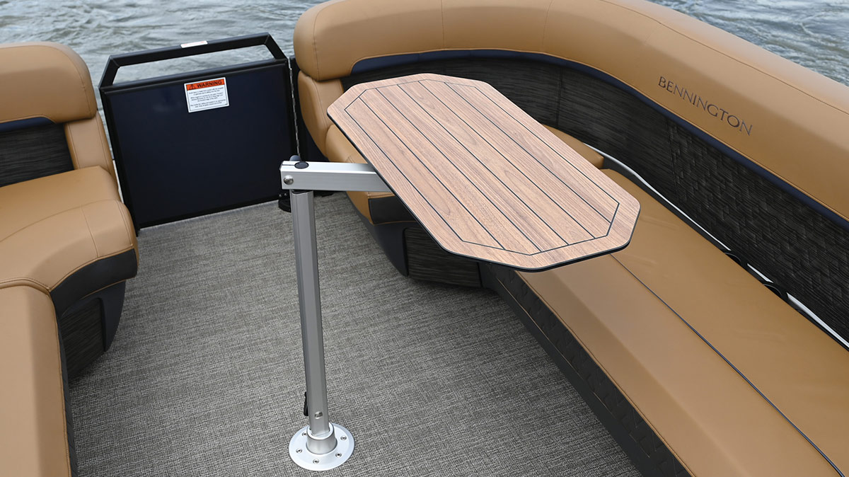 A wood-grain boat table mounted on an adjustable silver metal pedestal, installed on the grey deck of a pontoon boat surrounded by tan wrap-around Bennington seating.