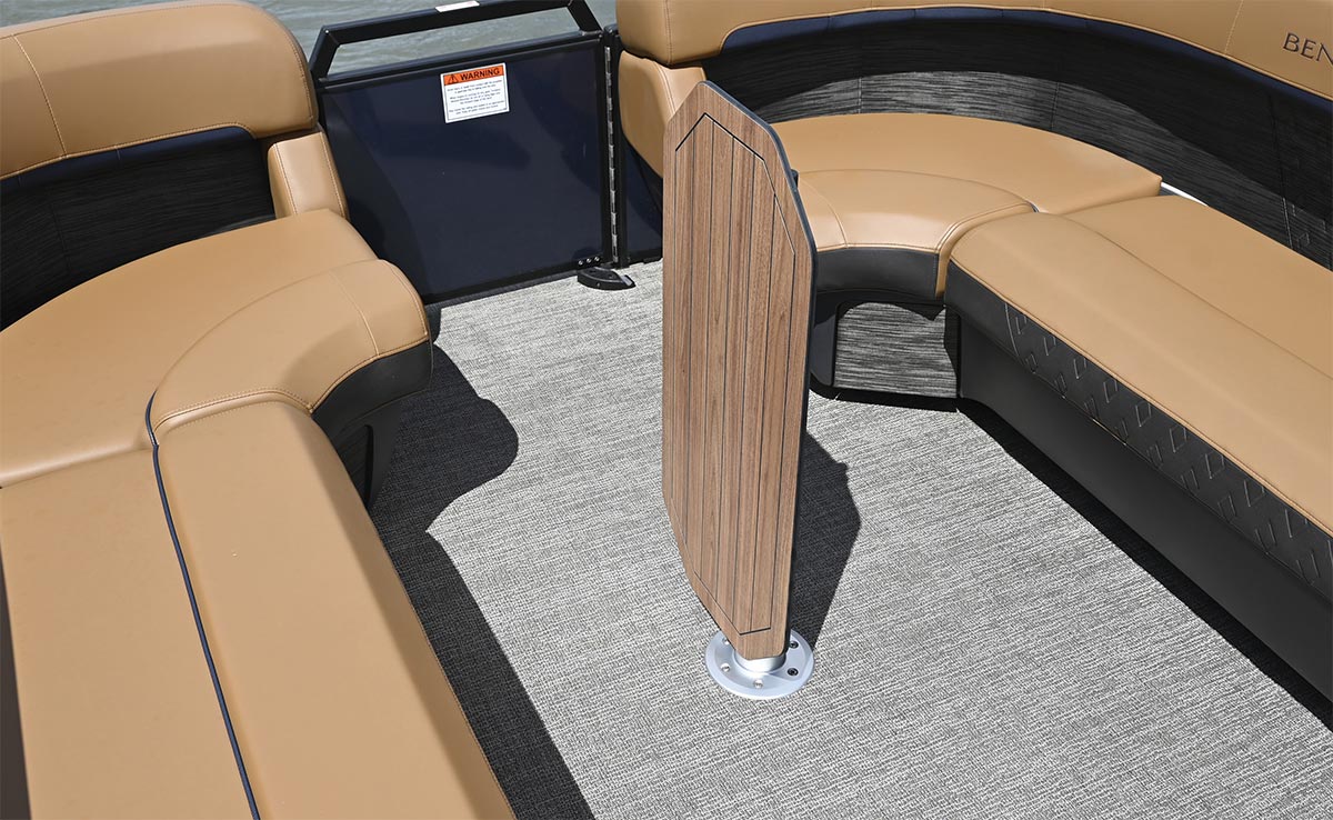 The wood-grain boat table is flipped vertically into a folded position on its silver metal pedestal, clearing the grey deck floor between tan wrap-around pontoon boat seats.