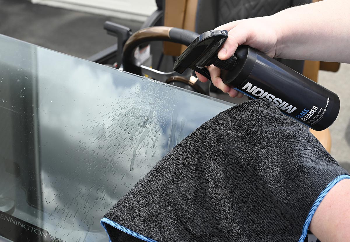 A close-up of hands spraying Mission brand Glass Cleaner onto a boat's windshield, with the other hand holding a dark grey microfiber towel ready to wipe the glass. The steering wheel is visible behind the windshield.