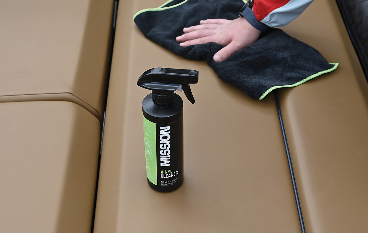 A black spray bottle of Mission Vinyl Cleaner sits on a tan boat seat while a person's hand uses a dark grey microfiber towel to clean the upholstery in the background.
